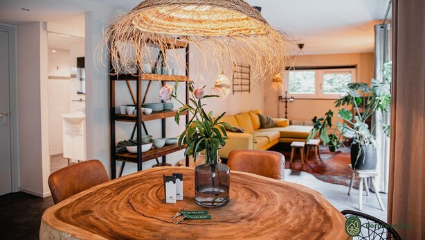 A dining area with a round wooden table, a woven hanging light fixture, and a vase with a plant.