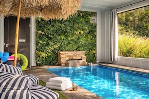 A pool with a stone wall and a waterfall feature, surrounded by a lush green wall and a thatched roof.
