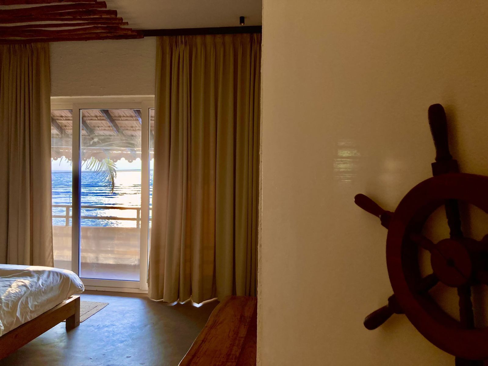 Una habitación con cama, un banco de madera, un adorno de volante y vista al océano por una puerta de vidrio.