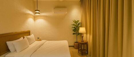A hotel room with a wooden bed, a small bedside table with a lamp, a potted plant, and a window with curtains.