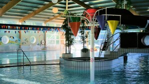 An indoor swimming pool with a water slide, surrounded by tiled walls and a ceiling with wooden beams.
