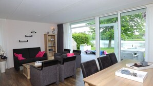 A modern living room with a sofa, armchairs, a coffee table, and a dining table. There is a bookshelf and a view of a lake through large windows.