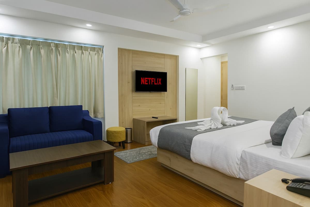 A modern hotel room with a blue sofa, a bed with white linens, a wooden nightstand, and a TV mounted on the wall displaying 'NETFLIX'.