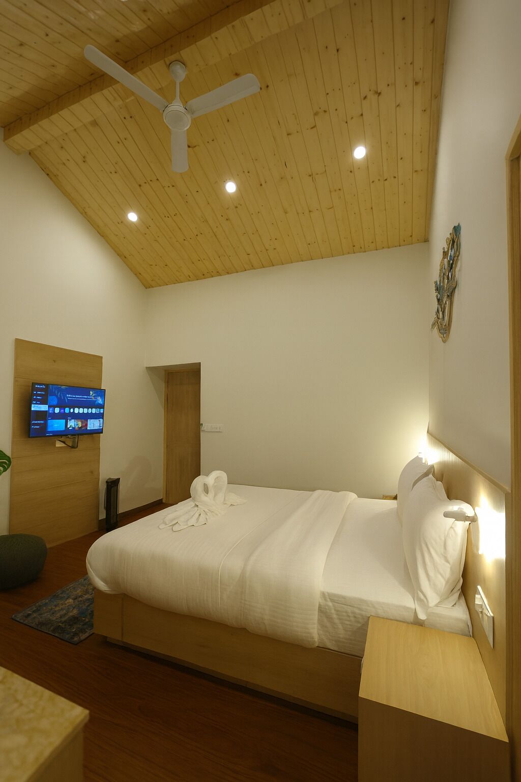A modern bedroom with a wooden ceiling, a bed with white linens, a TV on the wall, and a small bedside table.