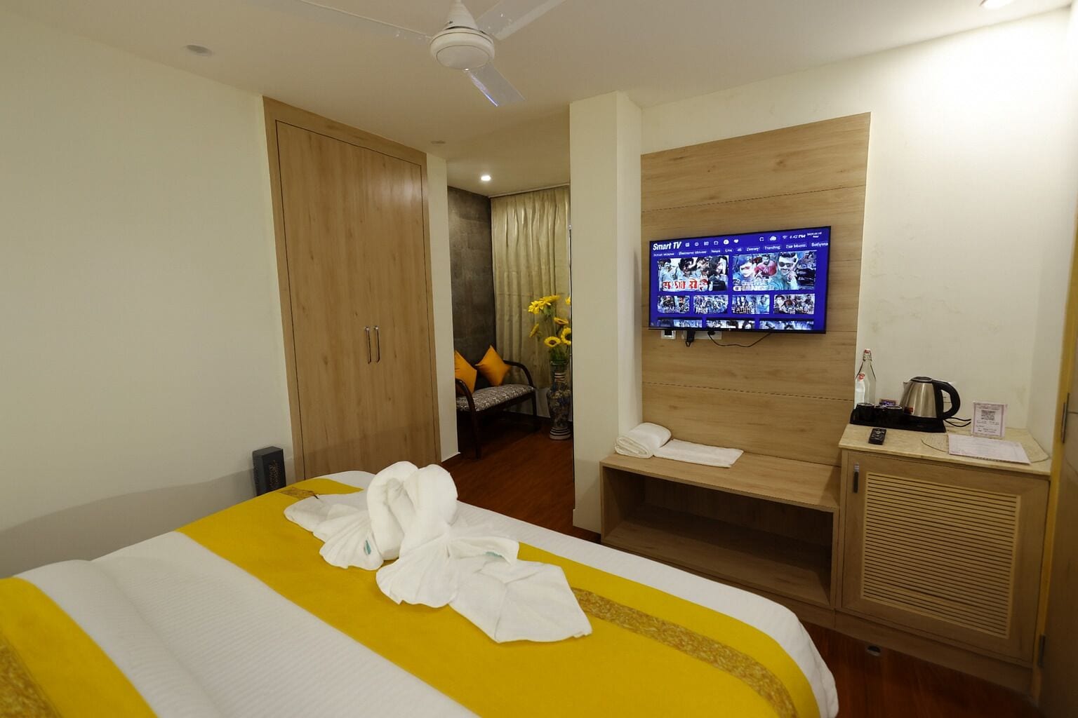 A hotel room with a bed, a TV mounted on the wall, a wooden wardrobe, a small table with a kettle, and a chair with a cushion.