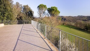 Zona exterior pavimentada amb barana metàl·lica, envoltada d'arbres i vista a turons ondulats.