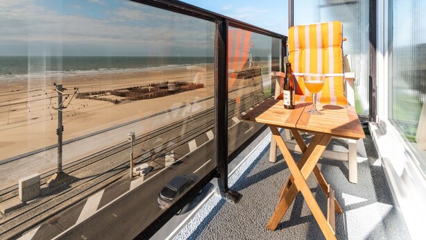 Un balcon donnant sur la plage, une table en bois avec une bouteille et un verre, et une chaise à rayures.