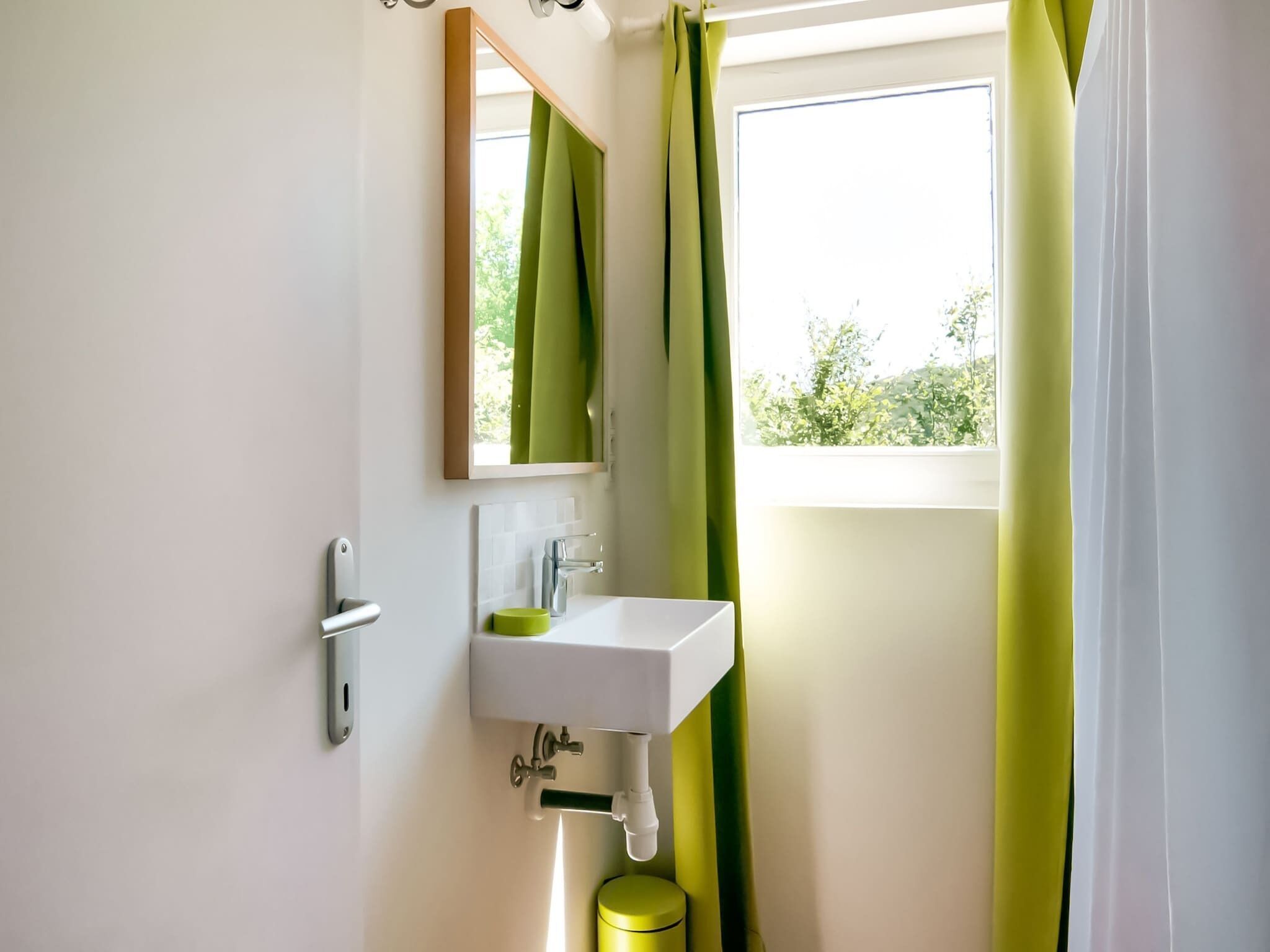 A bathroom with a white sink, a mirror, and a window with green curtains.
