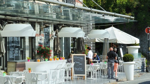 Un café en plein air avec des tables et des chaises blanches, un menu écrit à la craie et des plantes en pot.