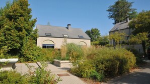 Une maison à deux étages, dotée d’un toit en pente, entourée d’une végétation luxuriante et d’arbres.