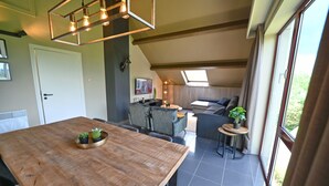 Une pièce spacieuse avec une table à manger en bois, un canapé et une petite table d’appoint sur laquelle est posée une plante. On aperçoit un luminaire au plafond et une porte au fond de la pièce.