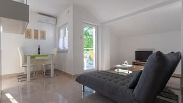 A modern living room with a grey sofa, a dining table with chairs, a TV, and a balcony with a view of greenery.