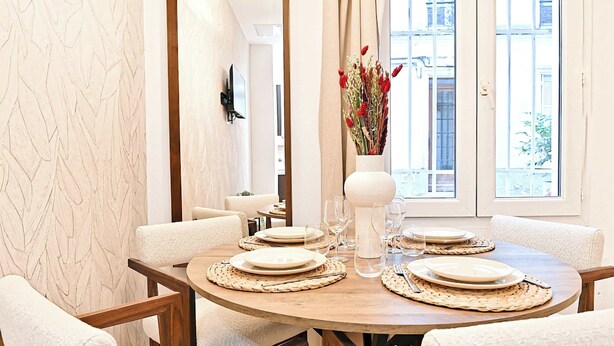 A round dining table with white plates, cutlery, and woven placemats, surrounded by white upholstered chairs. A vase with red flowers is on the table, and a large white vase is on the wall. - New flat with AC Porte Maillot/Arc de Triomphe (Paris)