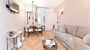 A modern living room with a herringbone floor, a sofa, a coffee table with a magazine, a dining area with a table and chairs, and a large window with curtains. - New flat with AC Porte Maillot/Arc de Triomphe (Paris)