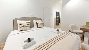 A neatly made bed with white linens and a beige throw blanket, a white chair, and a small wooden table with a green object on it. - New flat with AC Porte Maillot/Arc de Triomphe (Paris)