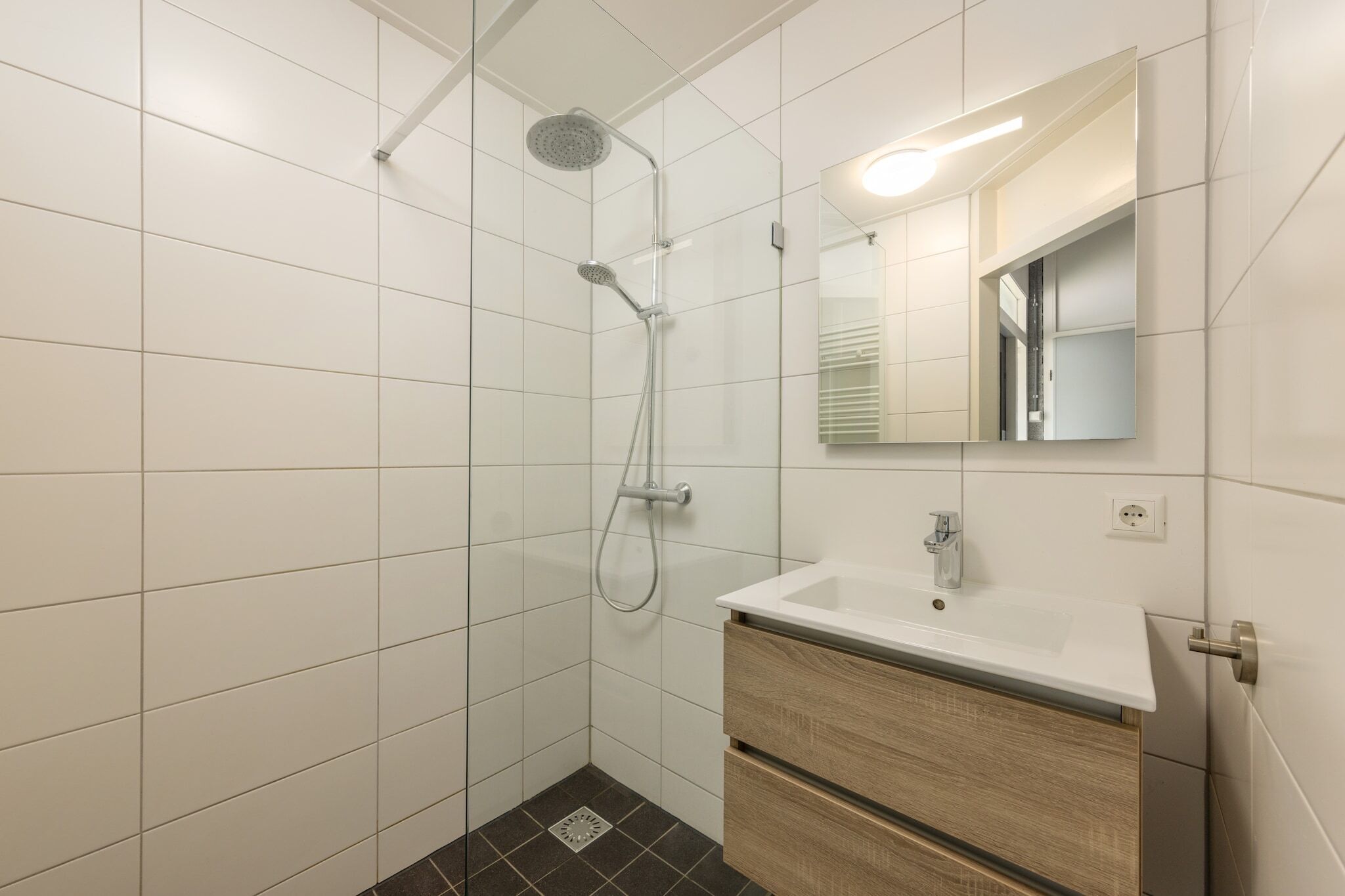 A modern bathroom with white tiles, a shower with a handheld showerhead, a sink with a mirror, and wooden cabinetry.