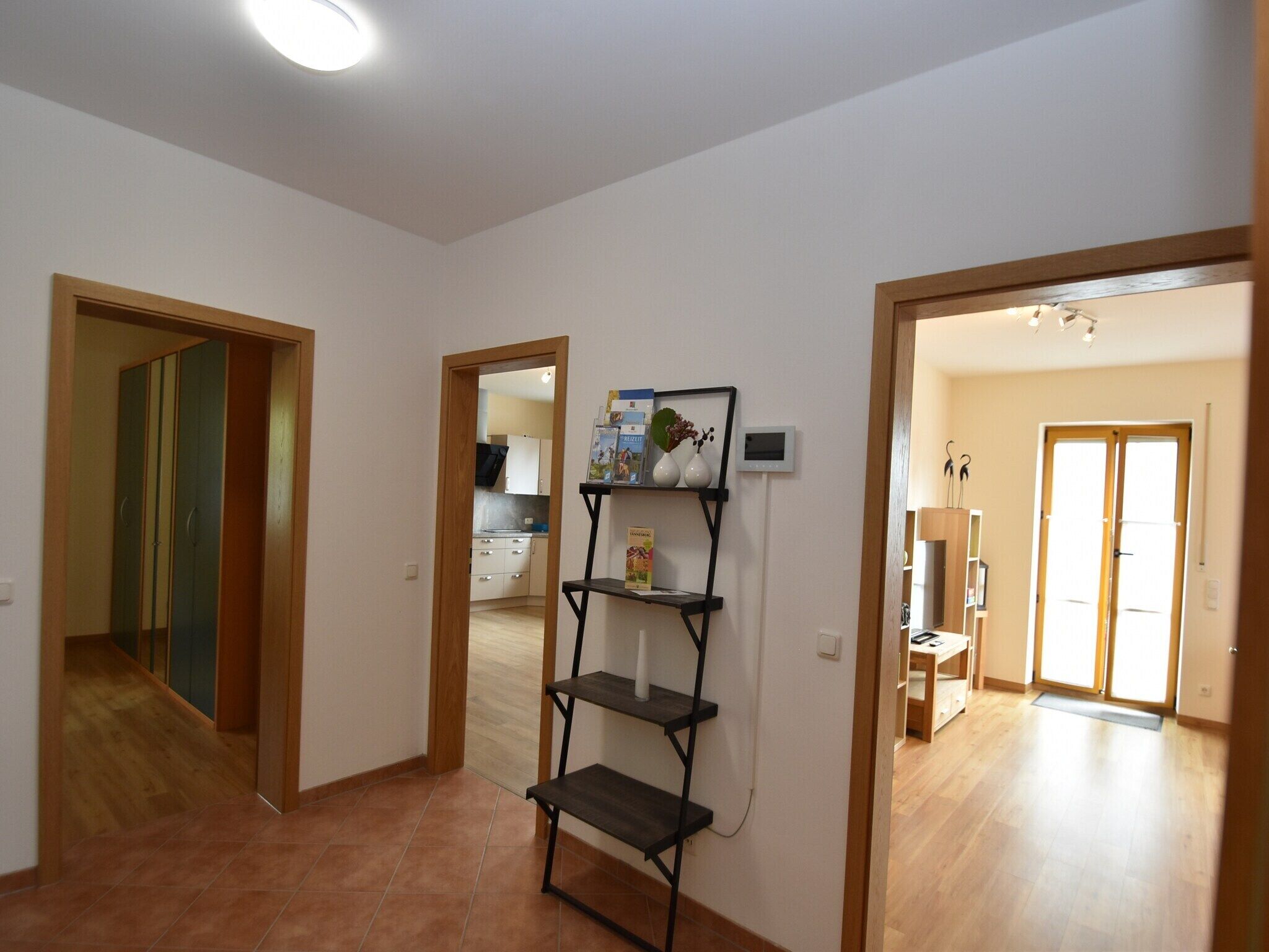 A spacious living area with a wooden floor, a black metal shelf unit, and a view into the kitchen through an open doorway.
