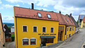 Un edificio amarillo con techo de tejas rojas, con un letrero que dice “CAFE KONDITOREI”. Tiene un escaparate con un toldo verde y dos ventanas adornadas con flores rojas.