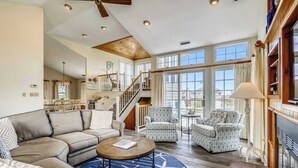 A spacious living room with a high ceiling, a staircase, and large windows. - Taylor, Ships Watch Community in Duck, NC (Kitty Hawk)