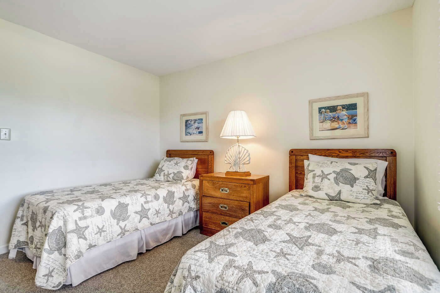 A room with two single beds, each with a wooden headboard and matching bedding. There is a nightstand with a lamp and a framed picture between the beds.