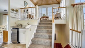 A staircase with a wooden finish, white railings, and a view of a dining area with wicker furniture. - Taylor, Ships Watch Community in Duck, NC (Kitty Hawk)