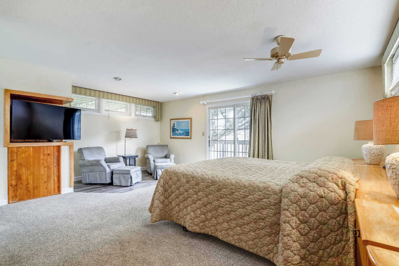 A hotel room with a bed, two armchairs, a TV, a ceiling fan, and a window with curtains.