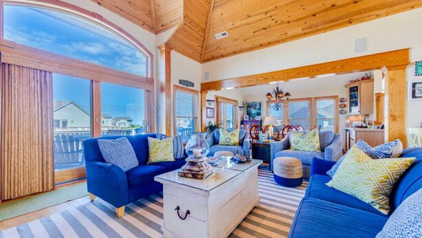A spacious living room with a large window, a blue sofa set, a white coffee table, and a kitchen area in the background. - Taylor, Ships Watch Community in Duck, NC (Kitty Hawk)