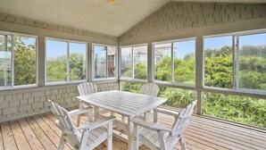 A covered patio with a table and chairs, large windows, and a view of greenery. - Taylor, Ships Watch Community in Duck, NC (Kitty Hawk)