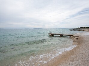 A beach with a wooden pier extending into the sea, clear water, and a distant shoreline with buildings. - Apartment on Pag near Seabeach and Town Center (Novalja)
