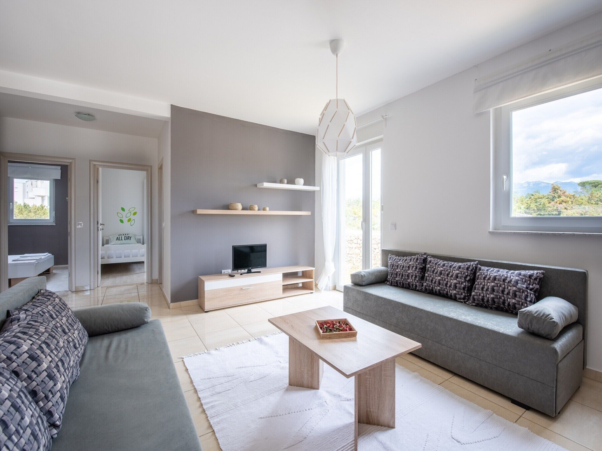 A modern living room with a grey sofa, a wooden coffee table, and a TV on a stand. There are shelves above the TV and a window with a view of the outdoors.
