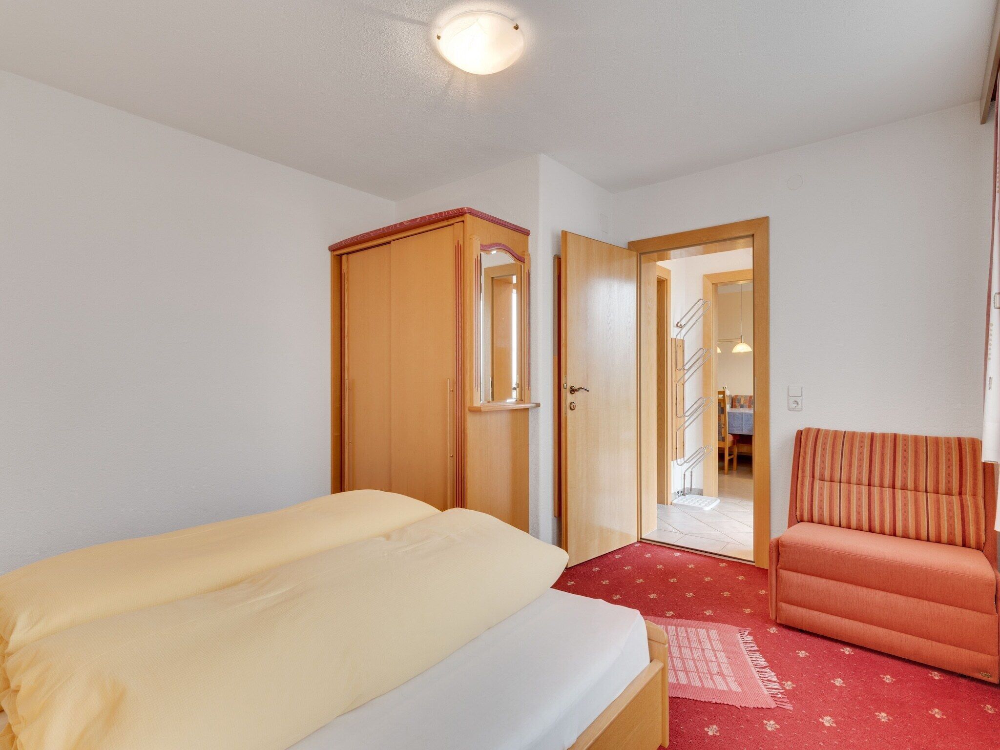 A bedroom with a bed, a chair, a wardrobe, and a door leading to a hallway.