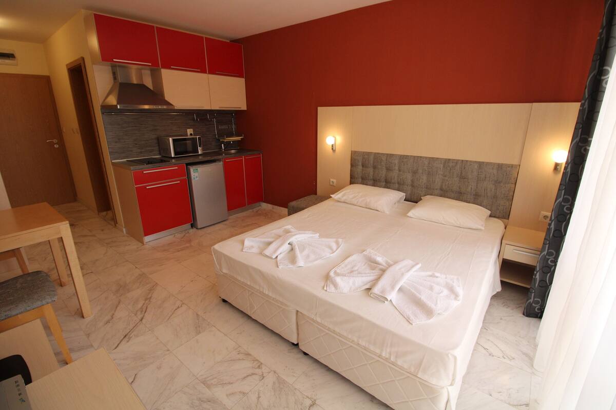 A hotel room with a bed, a desk, a kitchenette, and a red wall.