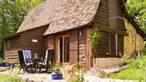 Un chalet rustique en bois, doté d’un toit en tuiles, d’un coin repas avec deux chaises et une table, et d’un jardin agrémenté de fleurs violettes.