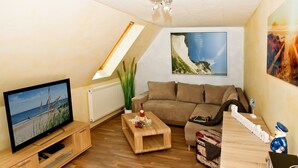 A cozy living room with a television, a sofa, a coffee table, and a small wooden table with a bottle and decorative items.