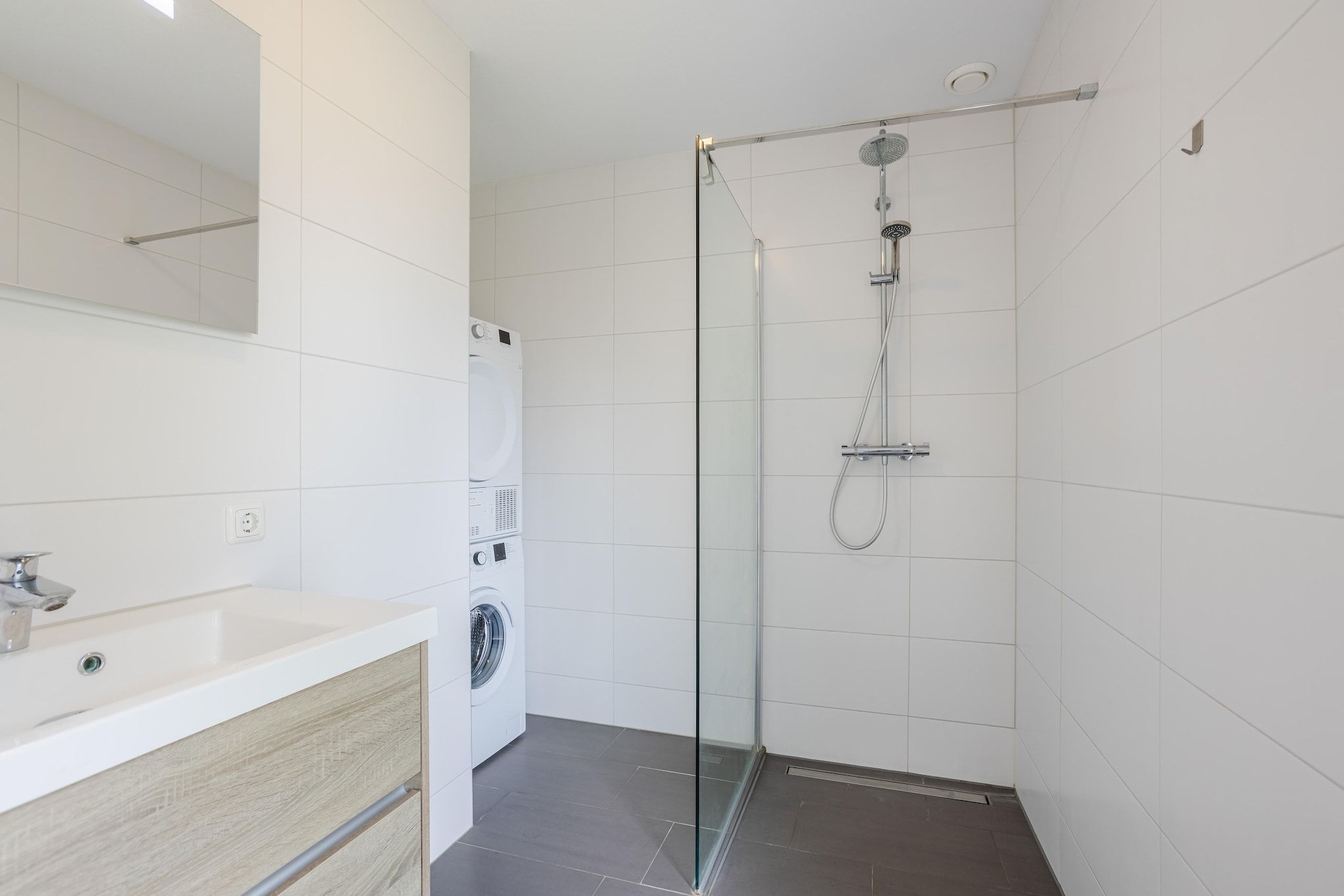 Un bagno moderno con doccia vetrata, lavatrice a incasso e lavandino bianco su un piano in legno.