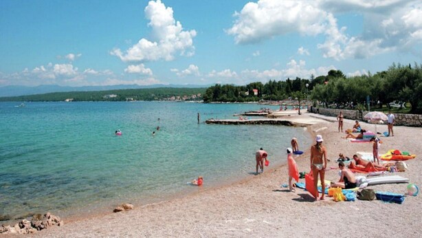 A beach with clear turquoise water, people swimming and sunbathing, and boats in the distance.
