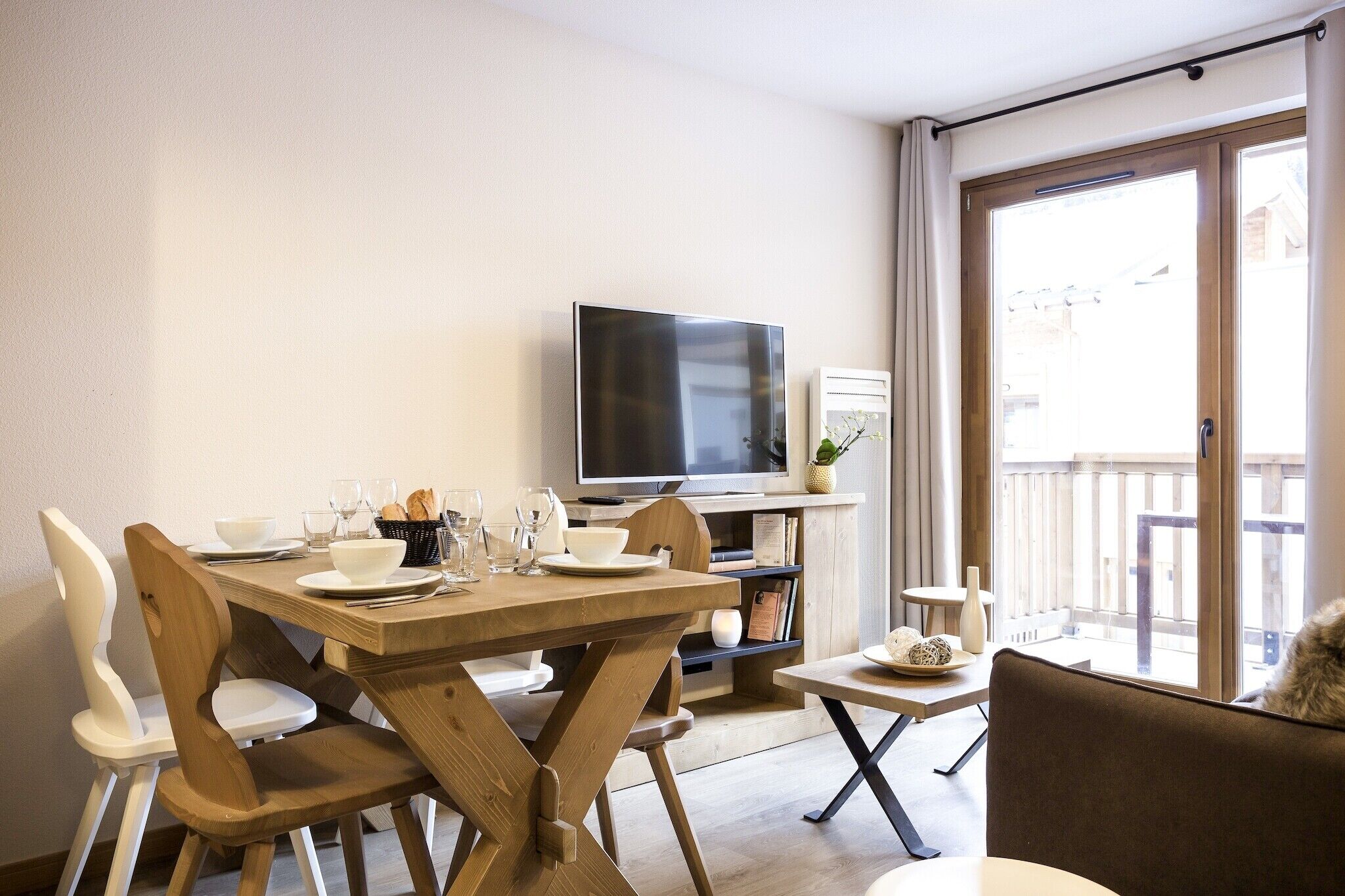 A dining area with a wooden table set for four, a television, and a view of the outside balcony.