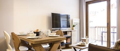 A dining area with a wooden table set for four, a television, and a view of the outside balcony.