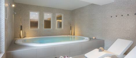 A modern spa area with a large bathtub, two lounge chairs, and a small table with toiletries.