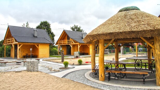 A wooden cabin with a thatched roof, a picnic area with benches, and a stone structure.