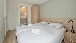 Ein modernes Hotelzimmer mit einem großen Bett, einem Holz-Kopfteil, einem Nachttisch mit Lampe und einem durch eine offene Tür einsehbaren Badezimmer.