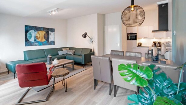 Ein modernes Wohnzimmer mit einer Couch, einem Esstisch und Stühlen. Im Hintergrund ist eine Küchenzeile zu sehen.