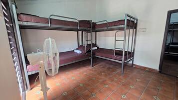 A dormitory room with bunk beds, a standing fan, and a tiled floor.