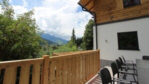 Una terraza de madera con vistas a montañas, un edificio blanco con una ventana y una mesa con sillas.
