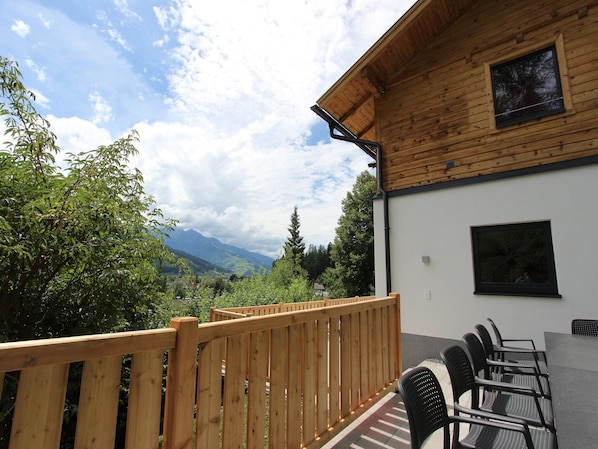 A wooden deck with a view of mountains, a white building with a window, and a table with chairs.
