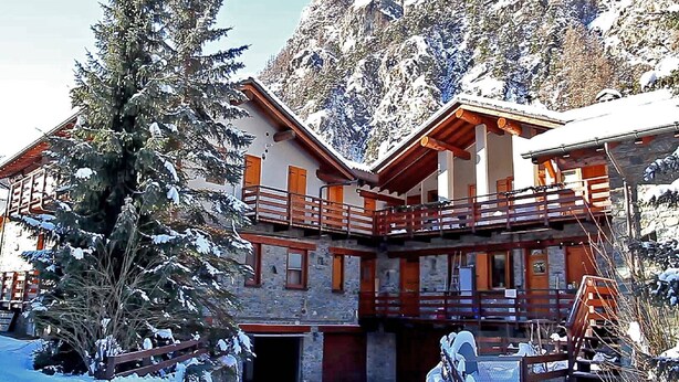Un bâtiment de style chalet, doté d’un balcon en bois, de murs en pierre et surplombant un paysage montagneux enneigé.
