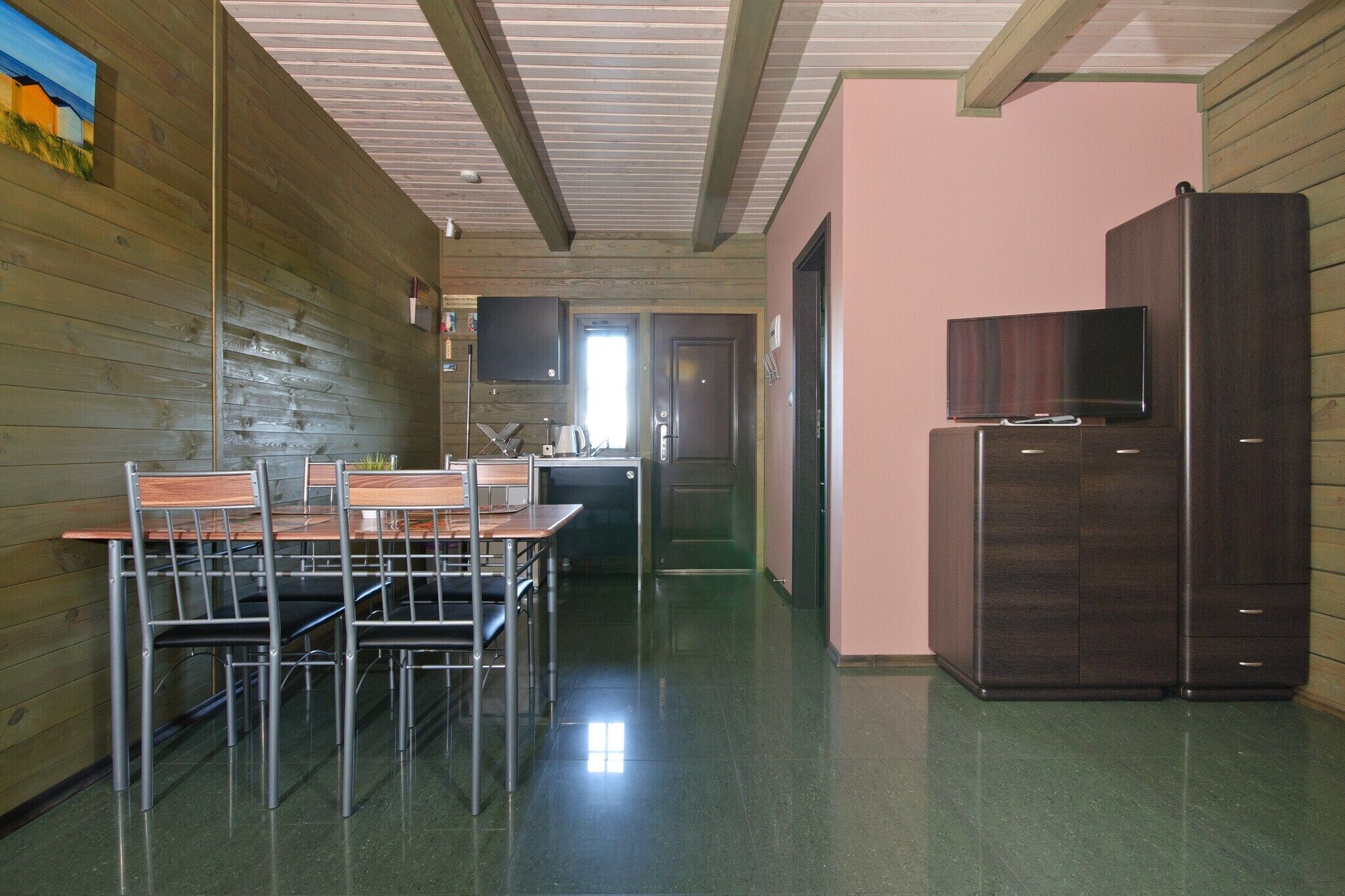 A room with a wooden wall, a dining table with chairs, a television on a cabinet, and a door leading to another room.