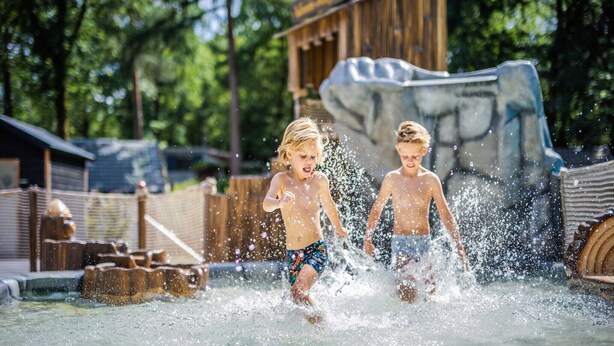 Dos niños jugando en una fuente de agua con una estructura de madera y una gran roca al fondo.