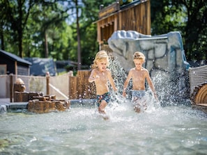 Two children playing in a water feature with a wooden structure and a large rock in the background.