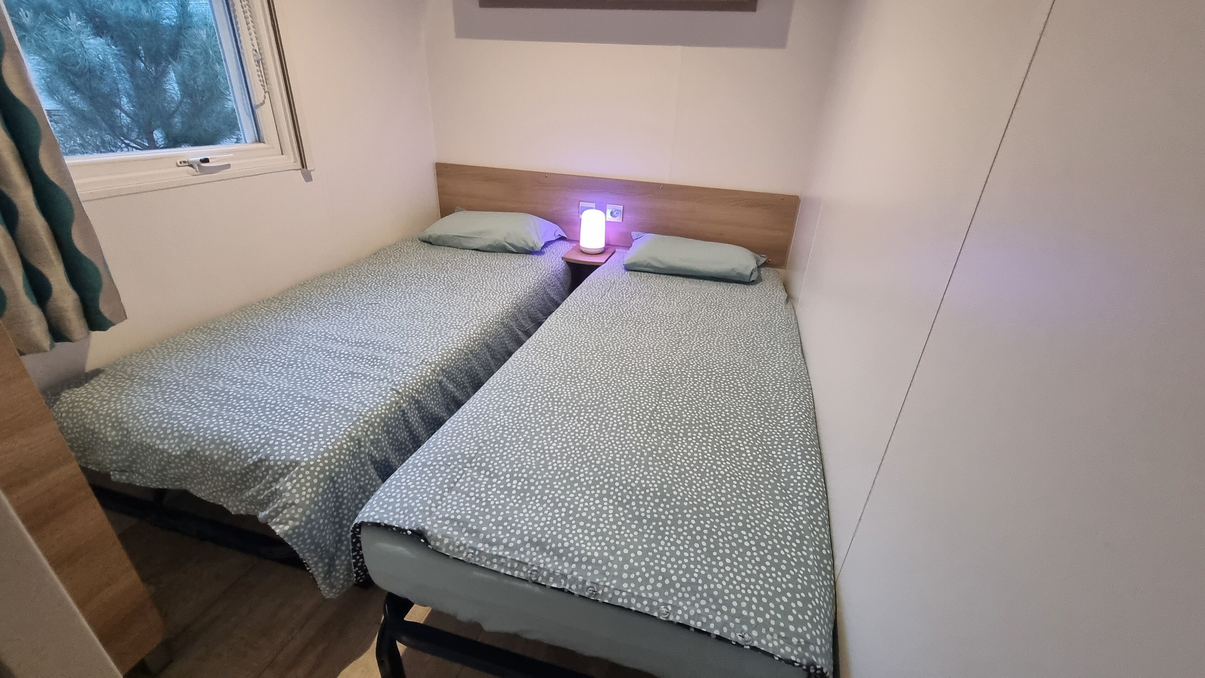 A small, single-room accommodation with two single beds, a nightstand with a lamp, and a window with a view of trees.